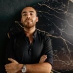 Portrait of a confident man with crossed arms in black attire against a marble background.